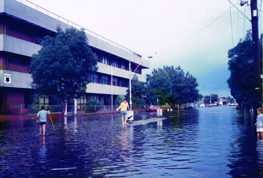 Water covers a road. A man stands next to a keep left sign on a traffic island in the middle. A young boy holding a stick wades through the water.