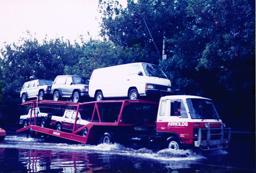 A truck carrying five vehicles drives on a flooded road. 