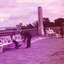 Unframed paintings are displayed on a paved path and on benches in front of a wall of hollow concrete blocks. A young girl sits on a bench watching a man who holds a painting. Two people look at paintings on another bench. Another person walks along the path. 