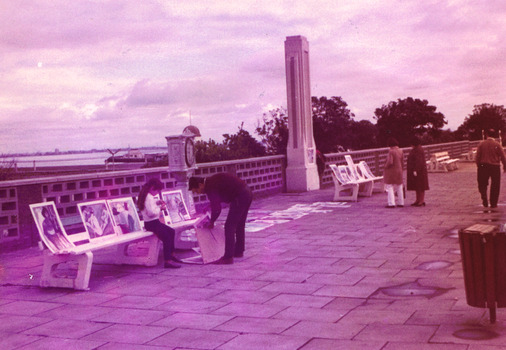 Unframed paintings are displayed on a paved path and on benches in front of a wall of hollow concrete blocks. A young girl sits on a bench watching a man who holds a painting. Two people look at paintings on another bench. Another person walks along the path. 
