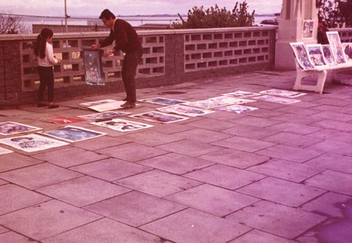 Unframed paintings are displayed on a paved path and on a bench in front of a wall of hollow concrete blocks. A man holds a painting that he is showing to a young girl.