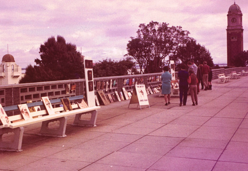 A row of benches along a paved path, in front of a wall of hollow concrete blocks. Unframed paintings are displayed along a bench and against the wall. Eight figures walk along the path and look at the paintings.