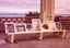Unframed paintings displayed along a bench and on a pillar on a paved path. Behind the bench is a wall of hollow concrete blocks through which can be seen the beach and sea in the distance. 