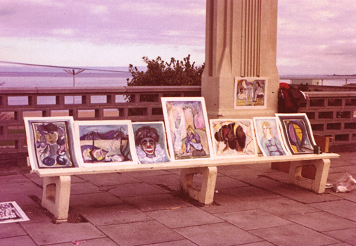Unframed paintings displayed along a bench and on a pillar on a paved path. Behind the bench is a wall of hollow concrete blocks through which can be seen the beach and sea in the distance. 