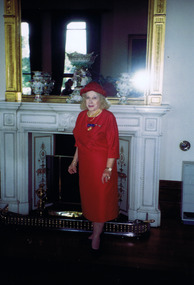 Woman in red dress and hat standing in front of ornate fireplace and mirror