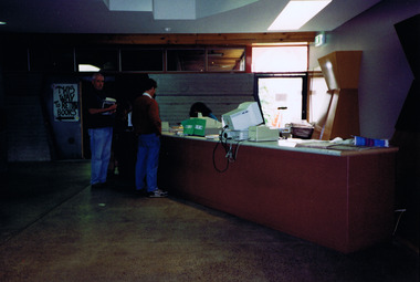 Two men stand at a desk in dimly lit room. A seated figure on the other side of the desk is partially obscured. Light enters through a window.