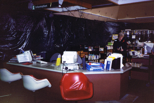 Three red and white swivel chairs in front of a desk and two blue chairs behind. On top of the desk are two computers and documents stored in binders. In the distance are trolleys with books on them and a woman who is standing while looking at books she is carrying.  Black plastic covers the walls .