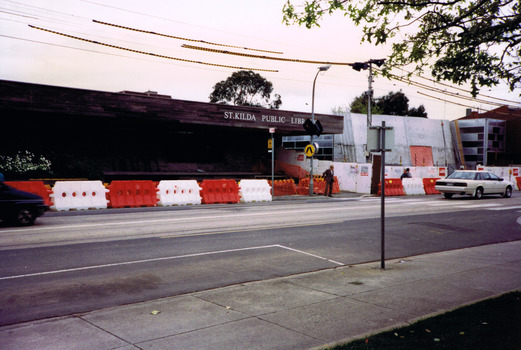 Red and white temporary safety barriers line an empty street in front of a building. Next to the building are vertical slabs of stone behind a temporary wooden fence and more safety barriers.