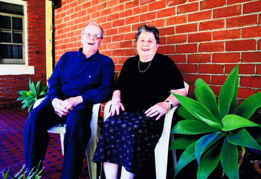 Two figures sitting on white chairs in front of a brick building.