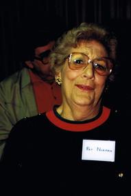 Head and shoulders of a woman wearing glasses and a black garment with a red neckline. A nametag is pinned to her clothing. A man is standing behind her in the background and is partly obscured.