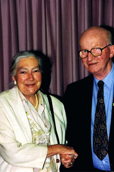 Two figures facing the camera. On the left is an elderly woman in a light coloured jacket and blouse. She is shaking hands with an elderly man wearing glasses, a blue shirt and a dark jacket.