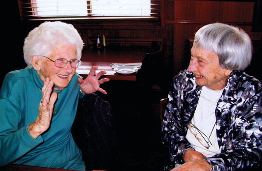 Head and shoulders of two smiling elderly women. Betty is on the left and is wearing glasses and a blue dress. The one on the right is wearing a white top and black and white patterned jacket. Behind them is a table set for a meal and a window with a venetian blind.