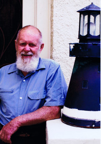 Head and shoulders of an elderly man in a blue shirt outside his house. Behind him is a white wall of the building. A blue light is on the right.