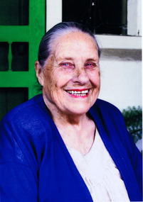 Head and shoulders of a smiling elderly woman in white blouse and blue cardigan in front of her house. Behind her is a green door, white wall, and stained glass window.