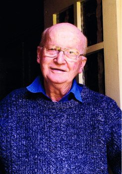 Head and shoulders of an elderly man wearing glasses, a blue shirt and blue jumper. He is standing in the open doorway of his house. Behind him is an yellow door with a paned glass window.