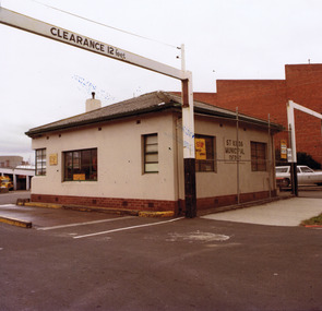 A bitumen driveway running along the side of a square white single storey brick building. In front of the building a wire fence and cement footpath. High over the driveway entrance is a beam, supported by two posts and painted with the words 'Clearance 12 feet'.