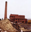 In the foreground a pile of rubble and rubbish. In the background a brick chimney and 3 storey brick building. 