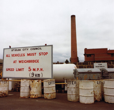 In the foreground a row of eight 44-gallon rusty metal drums in front of metal tanks. The drums are painted white and the word 'litter' is painted in black on 5 of them. The metal tanks are partly obscured by a sign that reads 'St Kilda City Council. All vehicles must stop at weighbridge. Speed limit 5 m.p.h. (9km)' . In the background a brick building and chimney. 