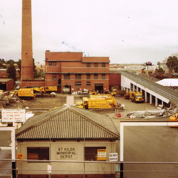A square white single storey building in the foreground is at the front of an open area. Five garbage trucks are in the open area. A road ramp on the right of the open area leads to a 3 storey brick building and chimney in the background.