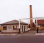 In the foreground a public street and footpath. Along the footpath a wire fence. Behind the fence, a square white single storey brick building. On the right of the building is a driveway. Signs on the fence on each side of the driveway say 'do not enter'. Over the driveway entrance is a high beam, supported by two posts. In the background is a brick chimney, another brick building, a van and garbage truck.