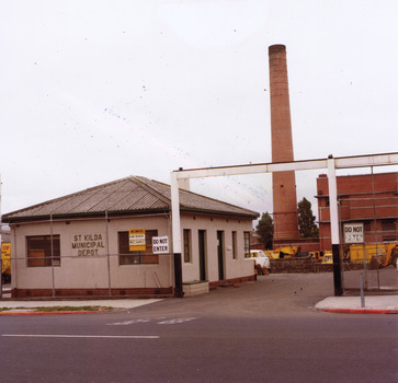 In the foreground a public street and footpath. Along the footpath a wire fence. Behind the fence, a square white single storey brick building. On the right of the building is a driveway. Signs on the fence on each side of the driveway say 'do not enter'. Over the driveway entrance is a high beam, supported by two posts. In the background is a brick chimney, another brick building, a van and garbage truck.