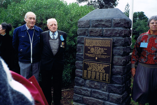 Three figures stand either side of a stone pillar. Two plaques are attached to the pillar.