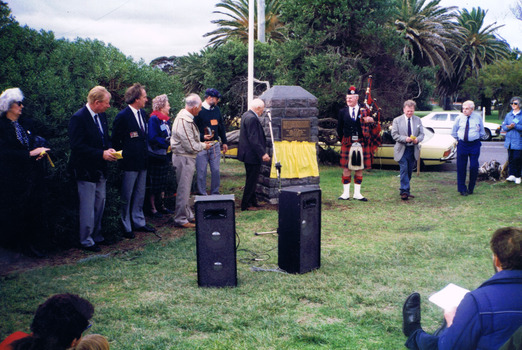 A crowd of people looks at a grey stone monument that has a yellow flag next to it. A man in the crowd is in Scottish highland dress and holds a set of bagpipes.