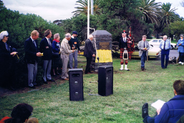 A crowd of people looks at a grey stone monument that has a yellow flag next to it. A man in the crowd is in Scottish highland dress and holds a set of bagpipes.