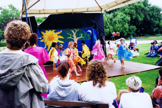 Girls in colourful costumes perform on a wooden stage, Other figures look on.