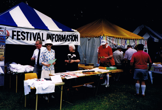 Three people stand behind a table at a gazebo that has a sign that reads "Festival Information". One man looks at a document taken from the table. Other people gather at another gazebo next to it.