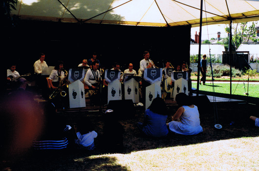 A band of musicians is playing brass instruments and an electric guitar on a stage in a marquee. A few figures are seated in front of them.