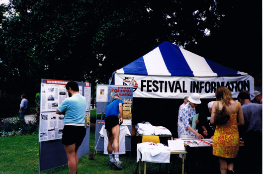 Figures look at items displayed at a gazebo that has a sign that reads "Festival Information"