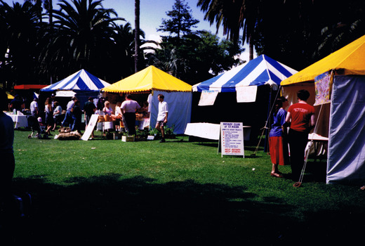 A row of colourful gazebos on a lawn. People on the lawn are visiting them.