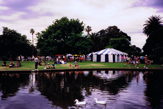 Figures sitting and standing on lawn near a pond. Two ducks swim in the pond. A striped canvas tent is on the lawn. 