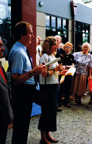 A woman addresses a group of people outside a building. A man stands next to her.