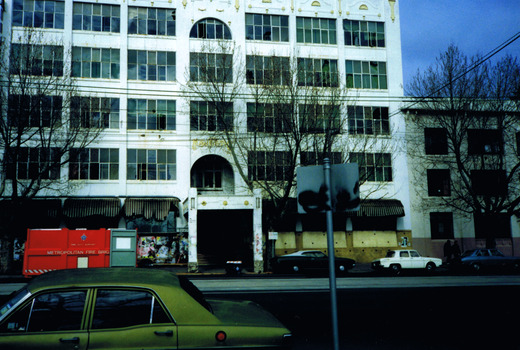 A multi-storey white building viewed from the opposite side of the street. A green parked car is in the foreground. Other cars are parked directly in front of the building. Also in front of the building is a large red container. On the side of the container the words "Fire duty support Metropolitan Fire Brig" are visible in yellow.
