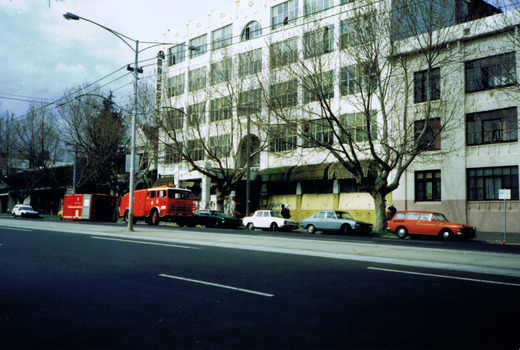 Multi-storey white building, seen at an angle from across a tree-lined street. Cars and a fire truck are parked out front. Also out front is a red container. 