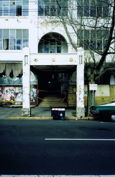 Entrance to multi-storey white building as viewed from the opposite side of the street. Wooden boards cover the building at ground level to the right of the entrance. The wall on the other side are covered with ripped posters. Awnings over the ground floor windows are ripped. 