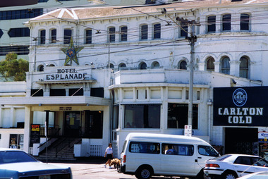 Multi-storey white building, with cars parked on the street in front. Near the cars a figure walks a dog. Wide stairs under a portico lead to the main entrance to the building. Above the portico is a sign that reads "Hotel Esplanade". Above the sign is a star. On the right of the image is a billboard advertisement for Carlton Gold beer. 