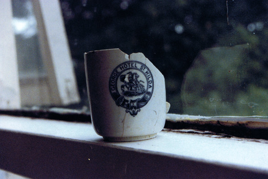 Side view of a white cup. It is cracked and missing the handle and almost half of the rim. It is decorated with a blue circular logo containing an image of a warrior on a horse and the words "George Hotel St Kilda"