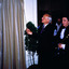 A man is facing a white wall that is partly obscured by a drawn curtain. He is holding a cord that will draw open the curtain. Another figure looks on.