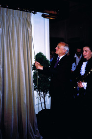 A man is facing a white wall that is partly obscured by a drawn curtain. He is holding a cord that will draw open the curtain. Another figure looks on.