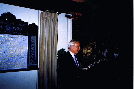 A man listening to a small group of people who are mainly obscured. On the wall behind him is a large plaque and a drawn curtain.