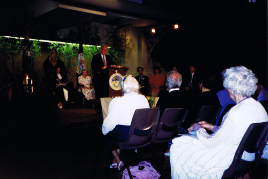 A man is standing on a stage behind a podium, addressing a group of people who are seated in front of him. Some people are also seated behind him on the stage. 