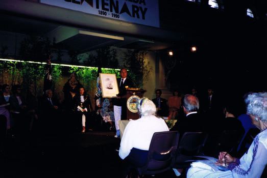 A man is standing on a stage behind a podium, holding a framed document. He is addressing a group of people who are seated in front of him. Some people are also seated behind him on the stage. 
