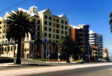 Six storey mustard-coloured building with multi-gabled roof on the corner of two streets. A high black veranda runs along the walls that face the streets. Tram tracks are in the foreground. Next to the building are older multi-storey apartment blocks.