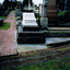 A grave in a graveyard. The headstone is the shape of a scroll and is mounted on a stack of three stone slabs. Words are inscribed on the headstone.