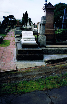 A grave in a graveyard. The headstone is the shape of a scroll and is mounted on a stack of three stone slabs. Words are inscribed on the headstone.