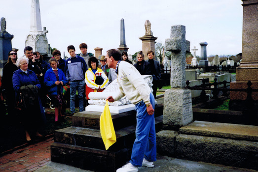 People in a graveyard watching a man remove a yellow veil from the base of a headstone of a grave. The headstone is in the shape of a scroll and is mounted on a stack of three stone slabs. 