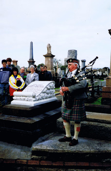 People in a graveyard listening to a man in a kilt who is standing next to a grave and playing bagpipes. The grave has a headstone, in the shape of a scroll, mounted on a stack of three stone slabs. 